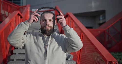 Bearded Man Puts On Headphones by Red Stairs