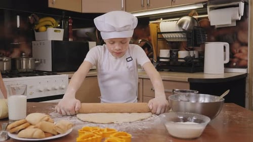 A Little Boy in an Apron and Cap Rolls Out Dough on a Table Using a Rolling Pin