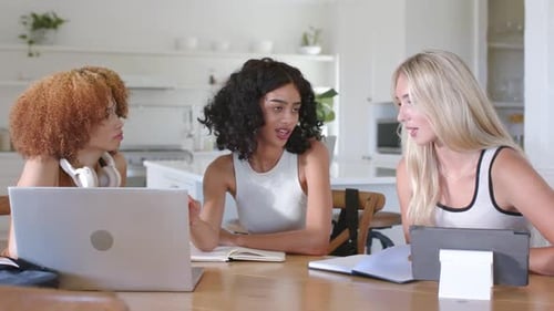 Trois femmes adultes assises à la table de la cuisine, femme centrale pointant vers un ordinateur portable, écrivant des notes sur une tâche