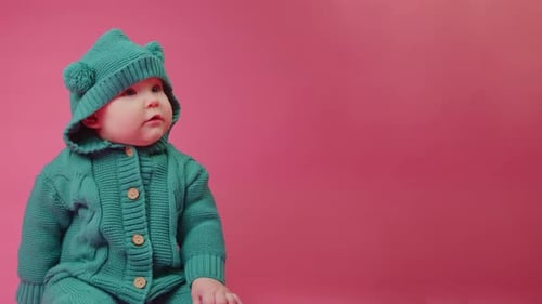 Portrait of a Cute Baby in a Warm Knitted Jumpsuit with a Hood on a Pink Background