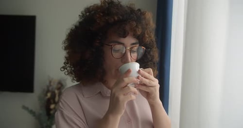 Beautiful Young Woman with Curly Hair Wearing Glasses Stands in Living Room By the Window Drinking
