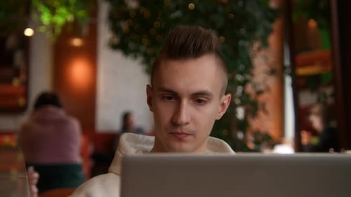 Young man is working on laptop in cafe and talking on phone. Stock footage. Young freelancer is work