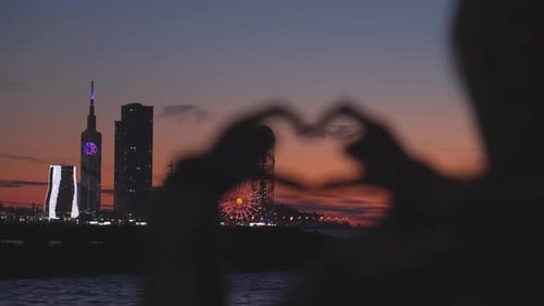 Mujer haciendo corazón con las manos con vista al brillante paisaje urbano al atardecer