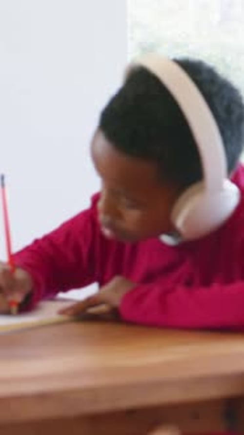 Vertical video: Placing hand on paper, kid in red shirt headset writing homework at table by window