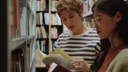 Two Female Students Discussing Books in Library