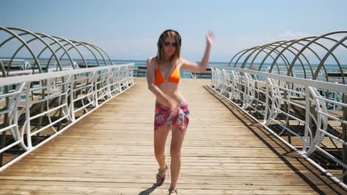 Young handsome woman in swim dress with headphones is dancing on wooden pier by sea water.