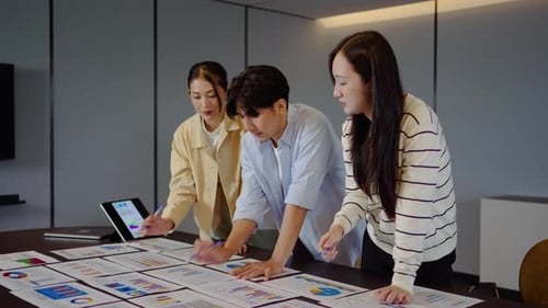 Asian business team analyzing financial data and charts on a desk in a corporate office meeting.