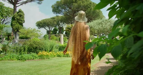 A woman walks through a manicured garden with tall trees and vibrant flowers in summer sunny day.