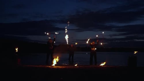 Group Of Professional Stunt Persons Performing Fire Show On Beach Of River In Summer Night