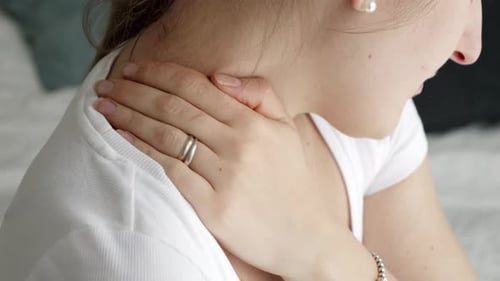 Nahaufnahme einer jungen Frau, die morgens auf dem Bett sitzt und sich den schmerzenden Hals reibt
