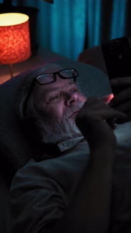 At Night Elderly Man with Glasses Lies in Bed and Surfing News on His Smartphone