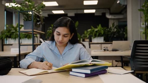 Focused Indian Student Writing in Notebook at Library Desk