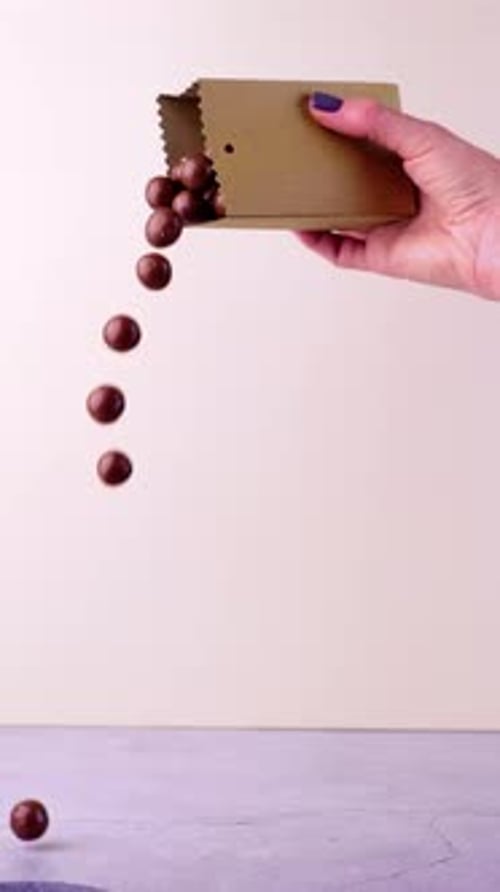 A close-up shot of a female hand holding a brown paper box, pouring balls in slow motion against a l