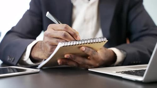 African American businessman writing in notebook while working on laptop in an office. Black man