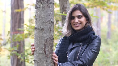 Brunette girl in the autumn forest stands by a tree and smiles at the camera