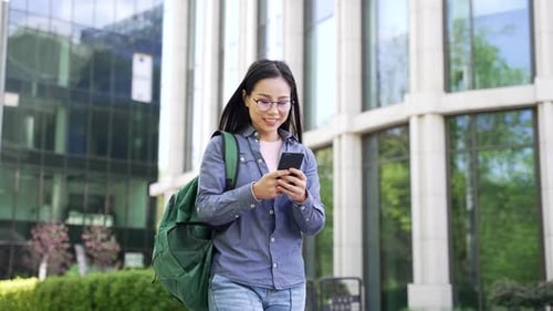 Smiling asian female student using phone walking on campus space near university building. A happy