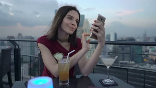Young woman texting on smartphone at rooftop bar in the city enjoying a cocktail