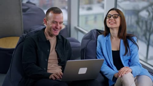 Positive pair of colleagues looking at screen of laptop, talking and laughing.