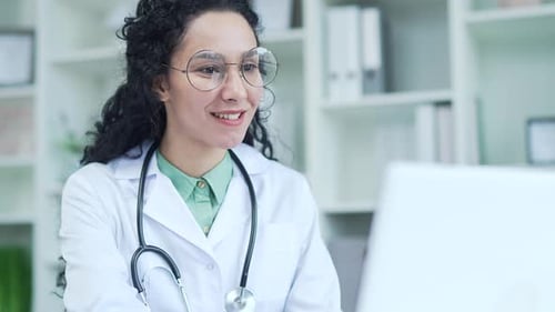 Close up. Smiling female doctor in a white coat writes a prescription looking at a laptop while sitt