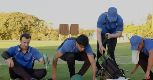 Diversos compañeros de equipo de críquet masculinos descansando con camisas azules en el campo, con un bate de cricket