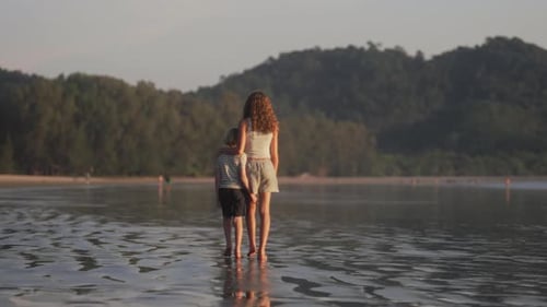 Young Siblings Strolling On Wet Beach Sand Against Lush Greenery