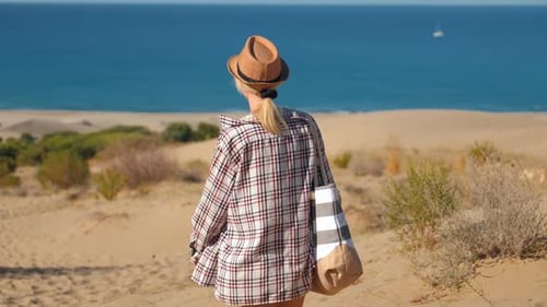 Young Woman Walking on Beach Towards Sea with Beach Bag and Hat