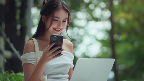 Happy asian woman in casual outfit working on laptop using mobile smartphone at urban park