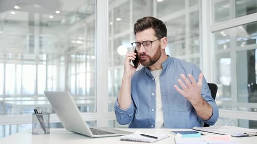 Angry businessman arguing talking on mobile phone sitting at workplace in office.