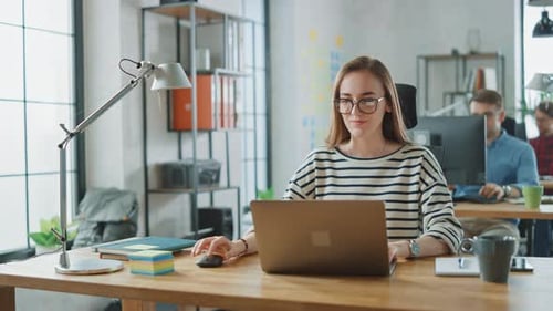 Beautiful Young Woman in Glasses is Working on a Laptop in a Creative Business Agency. They Work in