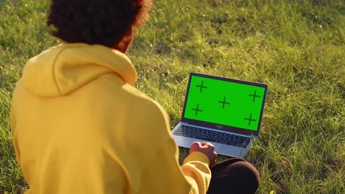 Young Guy in the Yellow Hoodie Sitting on the Grass in a City Park on a Sunny Day and Talking on a