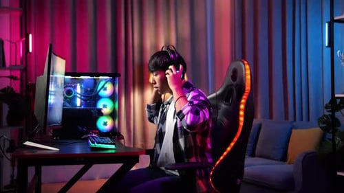 геймер в наушниках за компьютером с игрой на мониторе, атмосферное освещение
