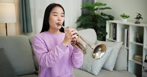 A young woman practices playing the trumpet while sitting on a sofa in a cozy living room.