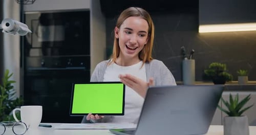 Hermosa joven sonriente que sostiene una tableta de pantalla verde mientras chatea en línea con sus socios.