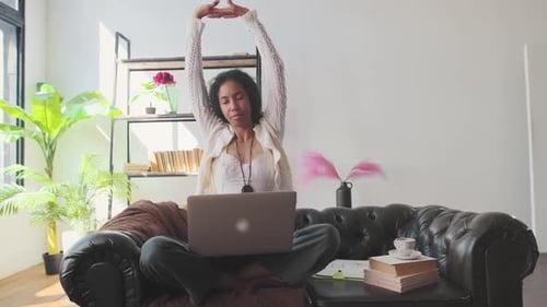 Young African American Woman Stretches Arms Up Sits on Sofa with Laptop on Lap