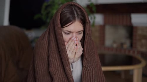Portrait, vue de face, d'une jeune femme triste dans une couverture, se frottant les mains, s'échauffant, assise à l'intérieur, malade