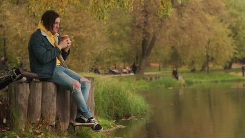 Young Handsome Man with Dreadlocks in Warm Clothes Drinking Coffee Near Lake in Golden Autumn