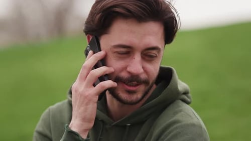 Bearded Brunette Man Talking on the Phone While Sitting on the Grass in the Park A Man is Talking on
