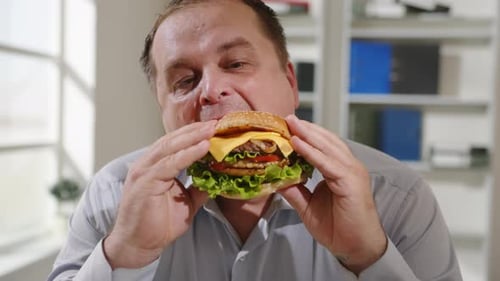 Close-up funny face man eating burger in office during lunch break.