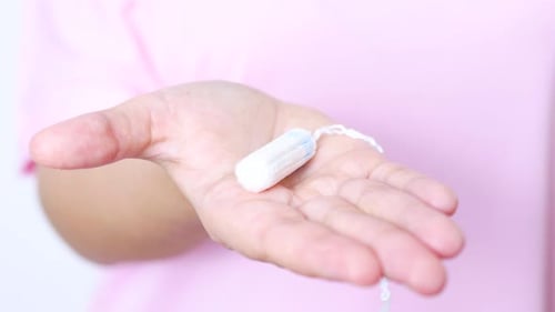 Frau präsentiert Damenhygiene-Produkt, ungeöffneter Tampon auf der Handfläche vor zartem Pastell