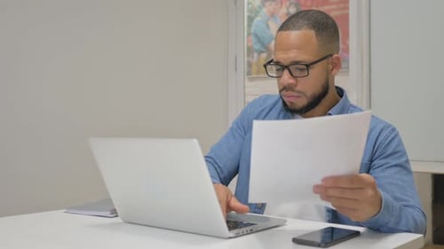 Man Reads Paper While Using Laptop At Desk