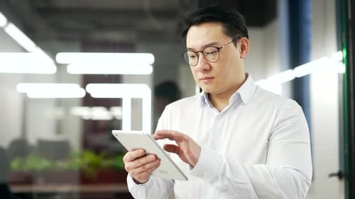 Serious asian businessman is using digital tablet while standing in business office. Focused man