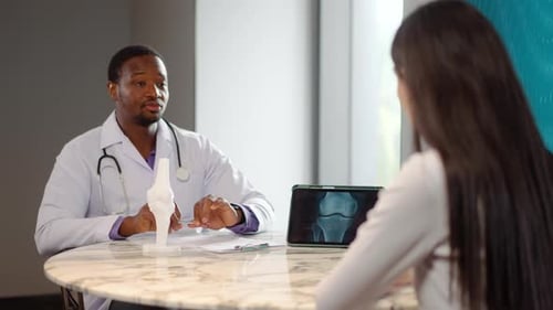 Männlicher Arzt befragt Patientin anhand eines anatomischen Modells und einer Röntgenaufnahme auf einem Tablet zu Knieschmerzen