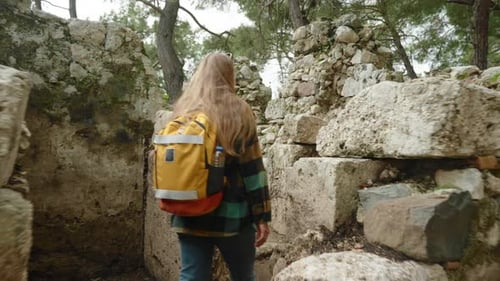 A woman with an orange backpack strolls through the ruined buildings of an ancient city.