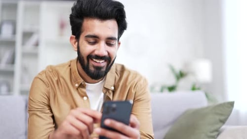 Close up. Young bearded man using smartphone sitting on sofa in living room at home.