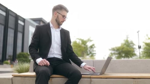 Focused Young Businessman Working on Laptop Outdoors in Modern Urban District