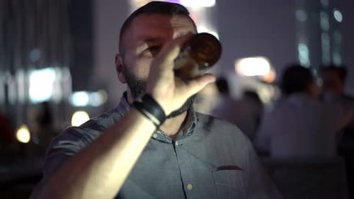 Happy young man enjoys beer at a luxury skybar overlooking the city at night