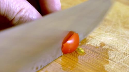 Male cutting a red cherry tomato with a very sharp silver knife on a wooden cutting board. Close up