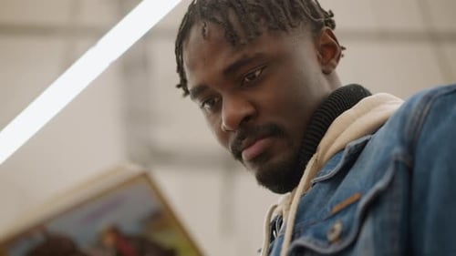 Male Focused on Reading Book Closely in Modern Library Surrounded By Colorful Wellorganized