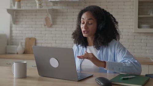 Femme parlant sur un appel en ligne depuis la cuisine portant des écouteurs et faisant des gestes