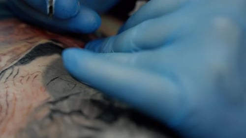Tätowierer mit blauen Handschuhen trägt mit einer Tattoo-Maschine Tinte auf die Haut eines Kunden auf. Der Vorgang